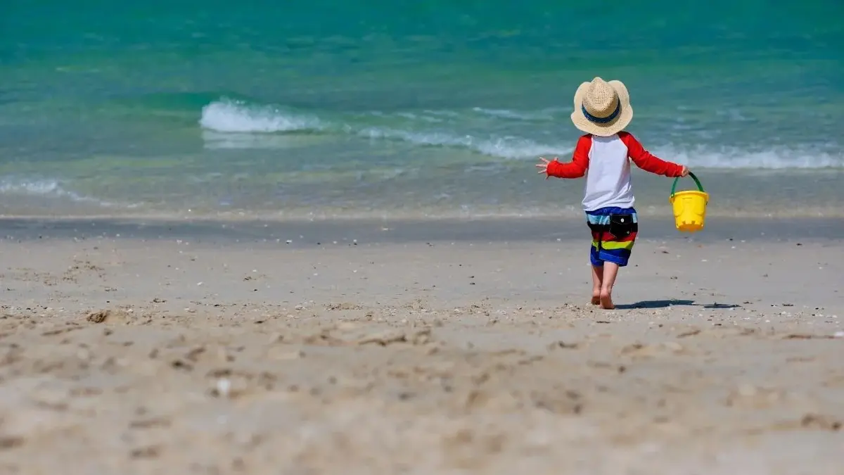 Dwuletnie dziecko w kapeluszu, z wiaderkiem, samo chodzi po plaży.