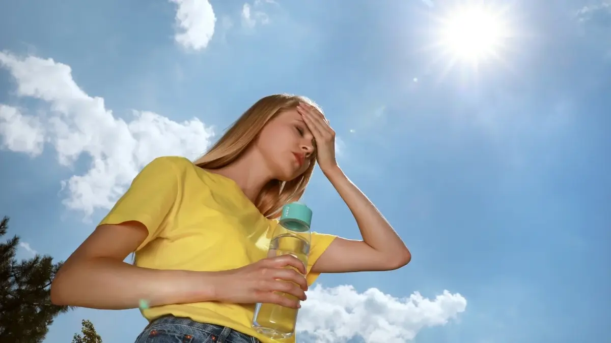 Kobieta w gorący dzień trzymająca się za głowę jedną ręką, w drugiej trzyma butelkę wody