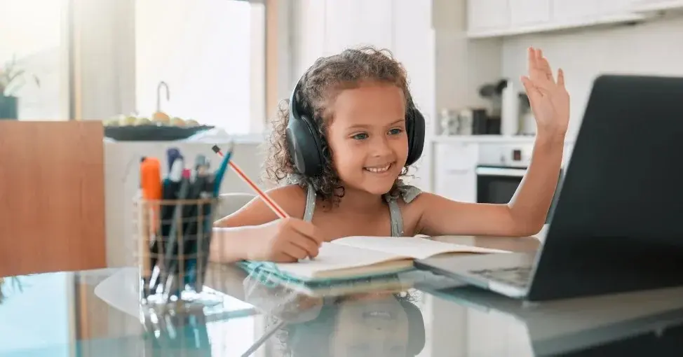 Przy biurku dla dziecka ze szklanym blatem siedzi uśmiechnięta dziewczynka w słuchawkach w trakcie zajęć przy komputerze