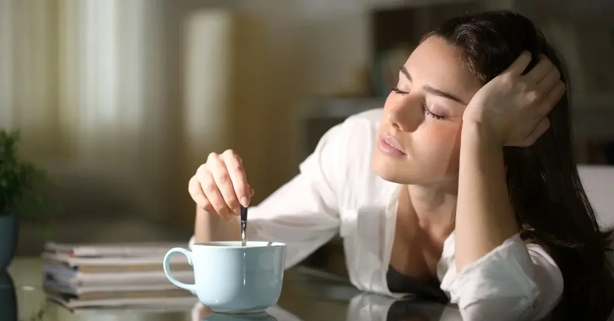 Zaspana kobieta z zamkniętymi oczami, miesza kawę w filiżance.