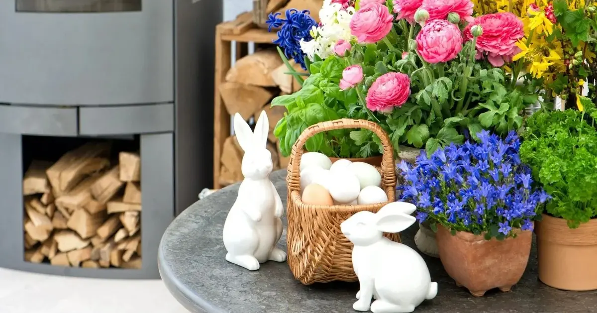 kwiaty kolorowe w doniczkach koszyk z jajkami figurki królików białe i kominek z drewienkami