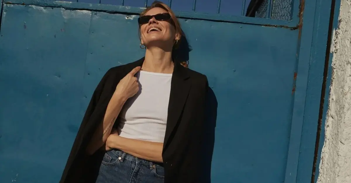 Uśmiechnięta kobieta w przeciwsłonecznych okularach w białym podkoszulku, czarnej marynarce i dżinsach 