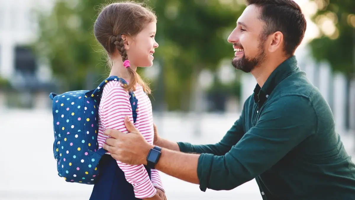 Mężczyzna żegna się z córką przed szkołą. Oboje się uśmiechają. Dziewczynka ma plecak i dwa warkocze.