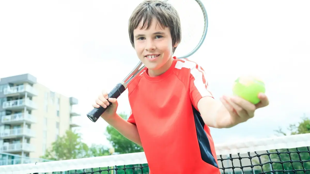 Chłopiec na korcie tenisowym trzymający w jednej ręce rakiete a w drugiej piłeczki do tenisa