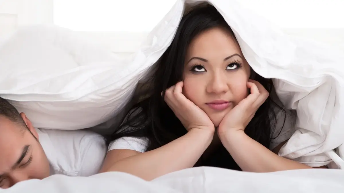 Kobieta leżąca w łóżku z poduszkami na głowie i twarzą opartą na dłoniach 