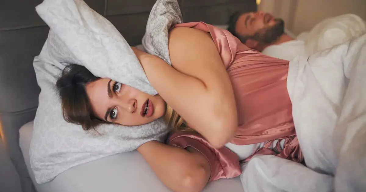 Kobieta w łóżku zasłaniająca głowę poduszką, w tle chrapiący mężczyzna