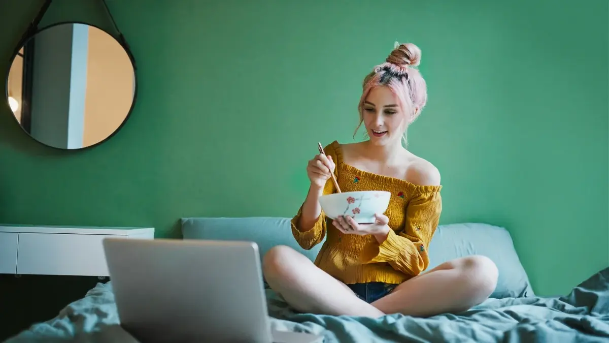 Kobieta siedząca na łóżku i jedząca coś z miski. Przed nią komputer