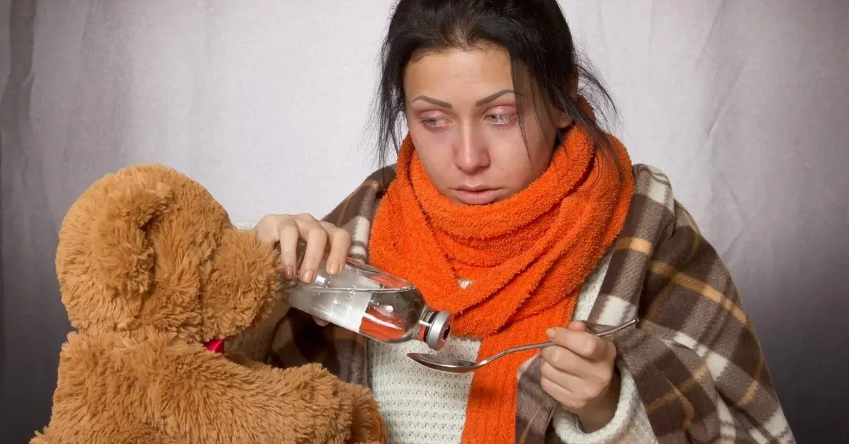 Kobieta w pomarańczowym szaliku nalewająca lek na łyżeczkę, obok pluszowy miś