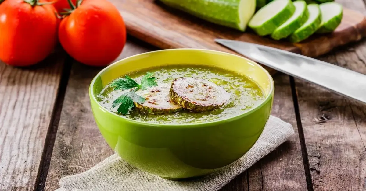 Zupa krem z cukinii w zielonej miseczce na drewnianym blacie. W tle pomidory, pokrojona cukinia i nóż