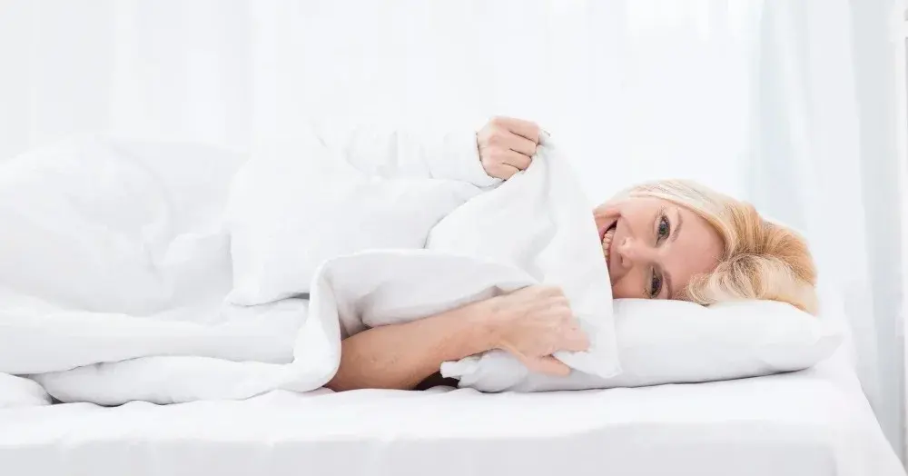 Kobieta leży na wygodnym materacu na łóżku w białej pościeli otulona kołdrą