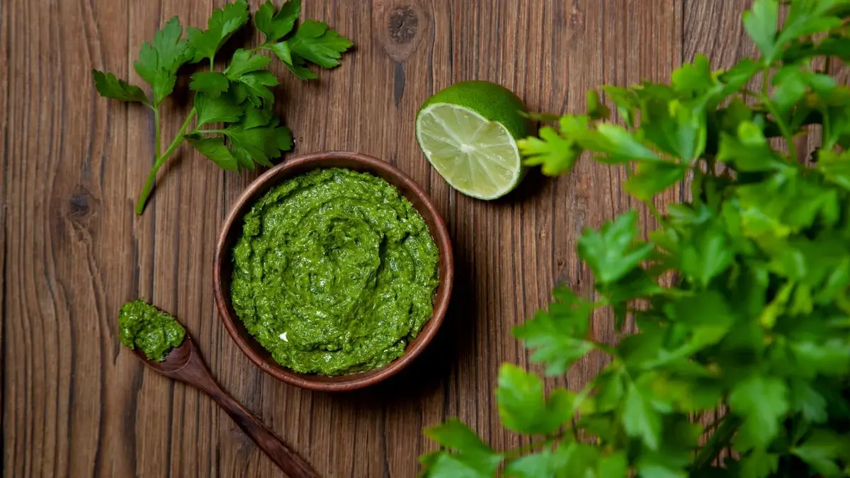 Na drewnianym blacie leży miseczka z zielonym pesto pietruszkowym. Obok limonka, łyżka u natka pietruszki.