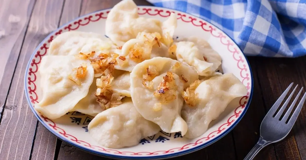 pierogi posypane cebulą w ozdobnym talerzu na drewnie obok ścierka w kratkę niebieska i widelec