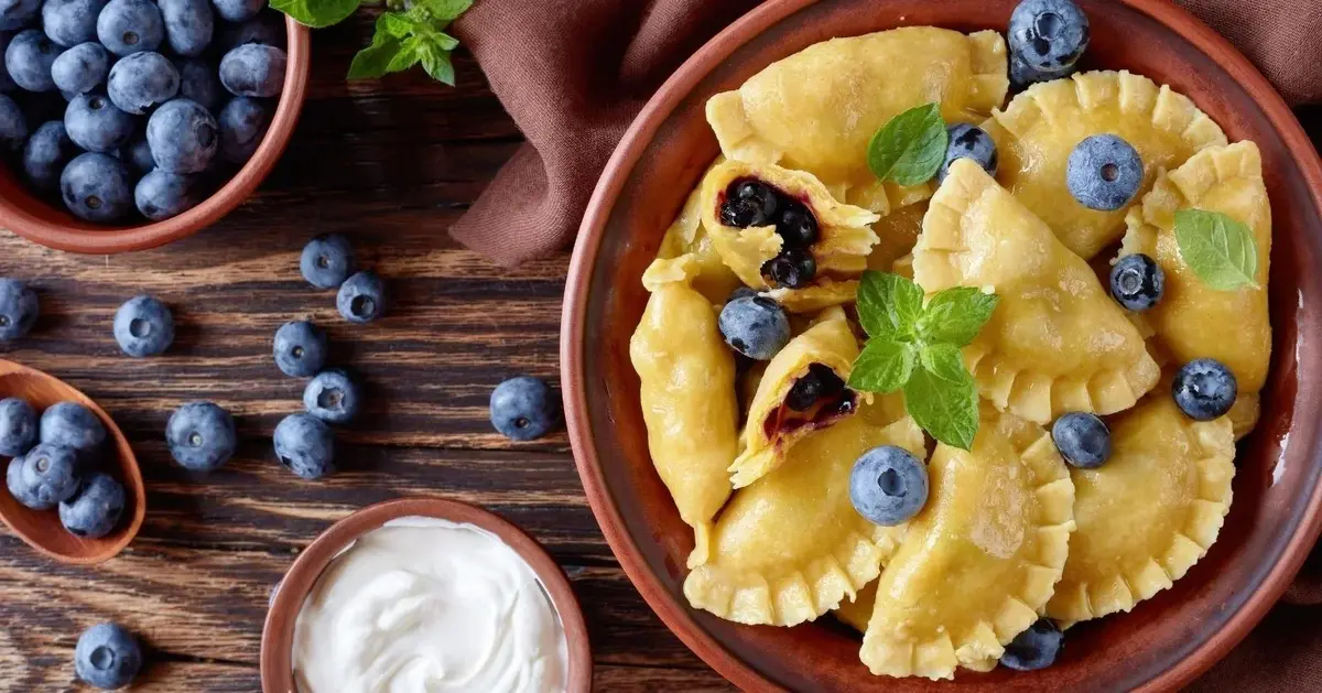 pierogi z jagodami na talerzu brązowym obok na tle drewna miska ze śietaną miska z borówkami i rozrzucone borówki