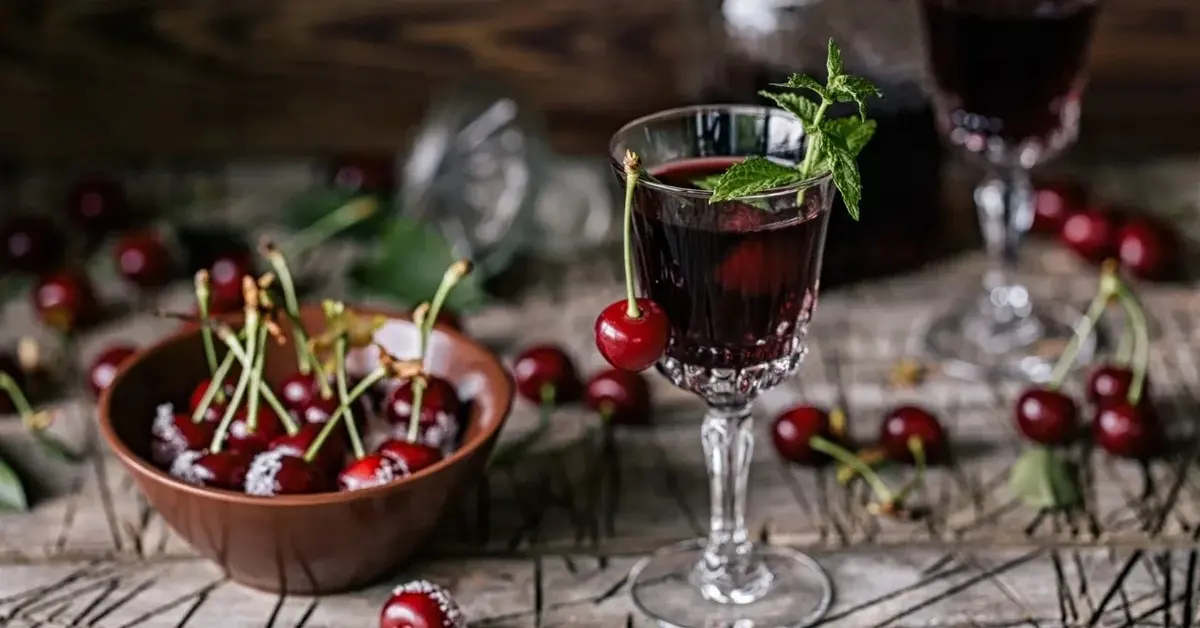 Nalewka z wiśni w kieliszku, obok miseczka z wiśniami i wiśnie poukładane dookoła