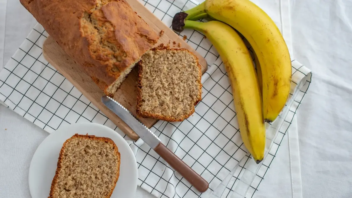 Ciasto bananowe na serwetce w kratke. Obok leżą dwa banany, dwa ukrojone kawałki ciasta i nóż