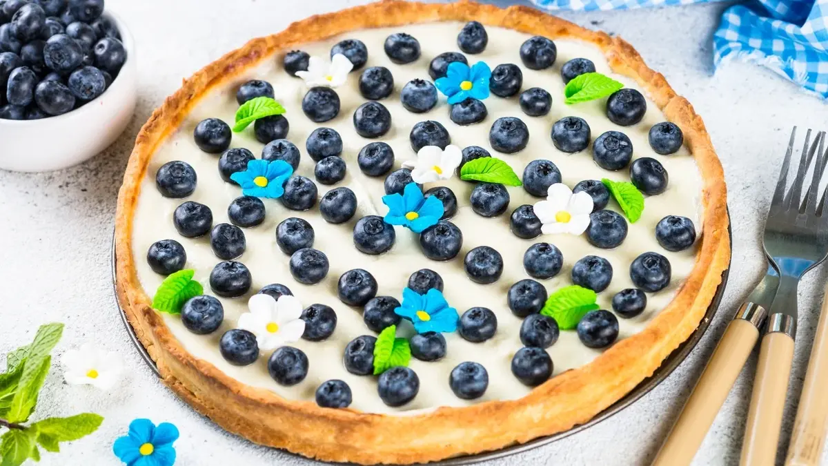 Okrągłe ciasto z mascarpone z kruchym brzegiem, kremem mascarpone  i borówkami. Obok nóż i widelec