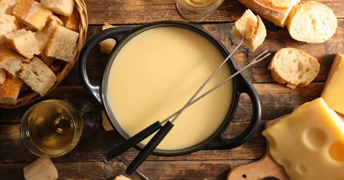 Fondue serowe w żeliwnym garnku, na górze dwa widelce. Dookoła kawałki bagietki