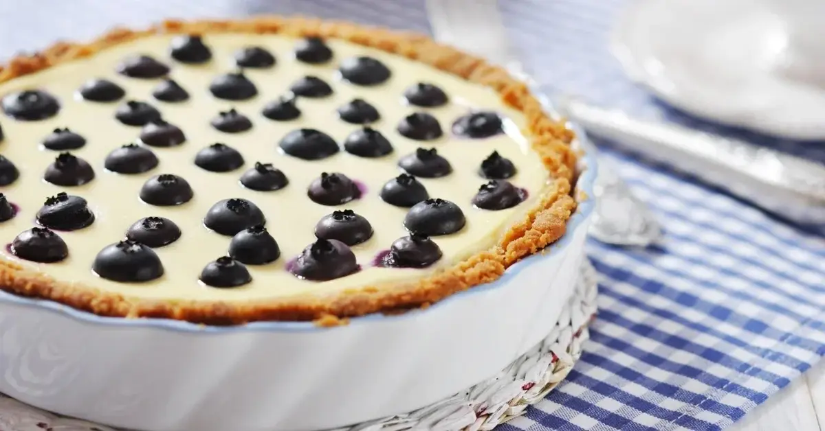 Tarta z borówkami w białym naczyniu do zapiekania stojącym na ściereczce w drobną niebiesko-białą kratkę