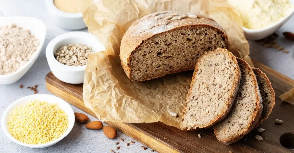 Bochenek bezglutenowego chleba i dwie kromki na desce drewnianej. Obok miseczki z różnymi mąkami