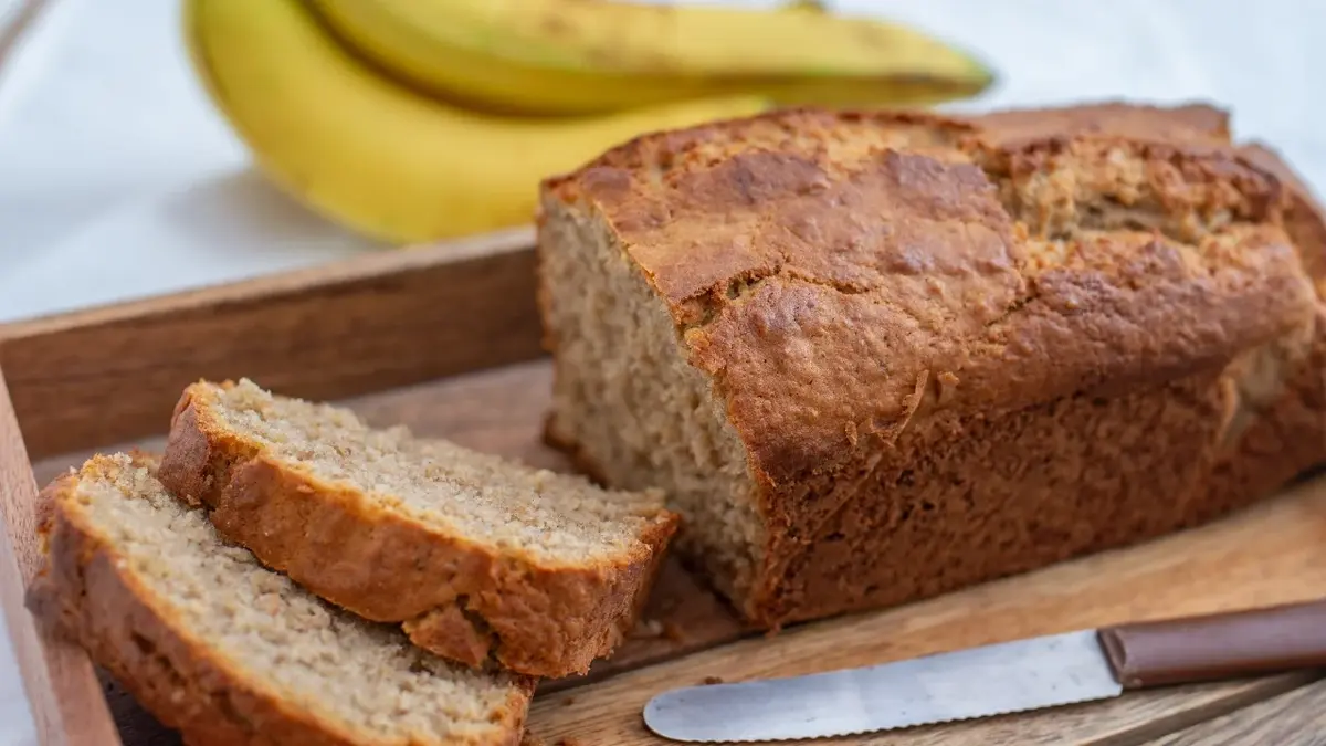 Chleb bananowy na drewnianej desce. Ukrojone dwa kawałki ciasta, w tle banany 