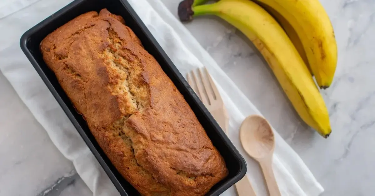 Chleb bananowy w formie do pieczenia na białej ściereczce. Obok dwa banany i drewniana łyżka i widelec