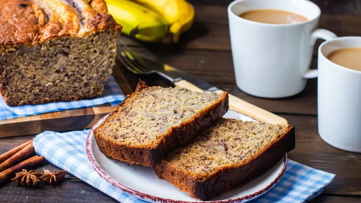 Chleb bananowy - dwa kawałki ciasta na talerzyku deserowym. W tle ciasto  kubeczki z kawą