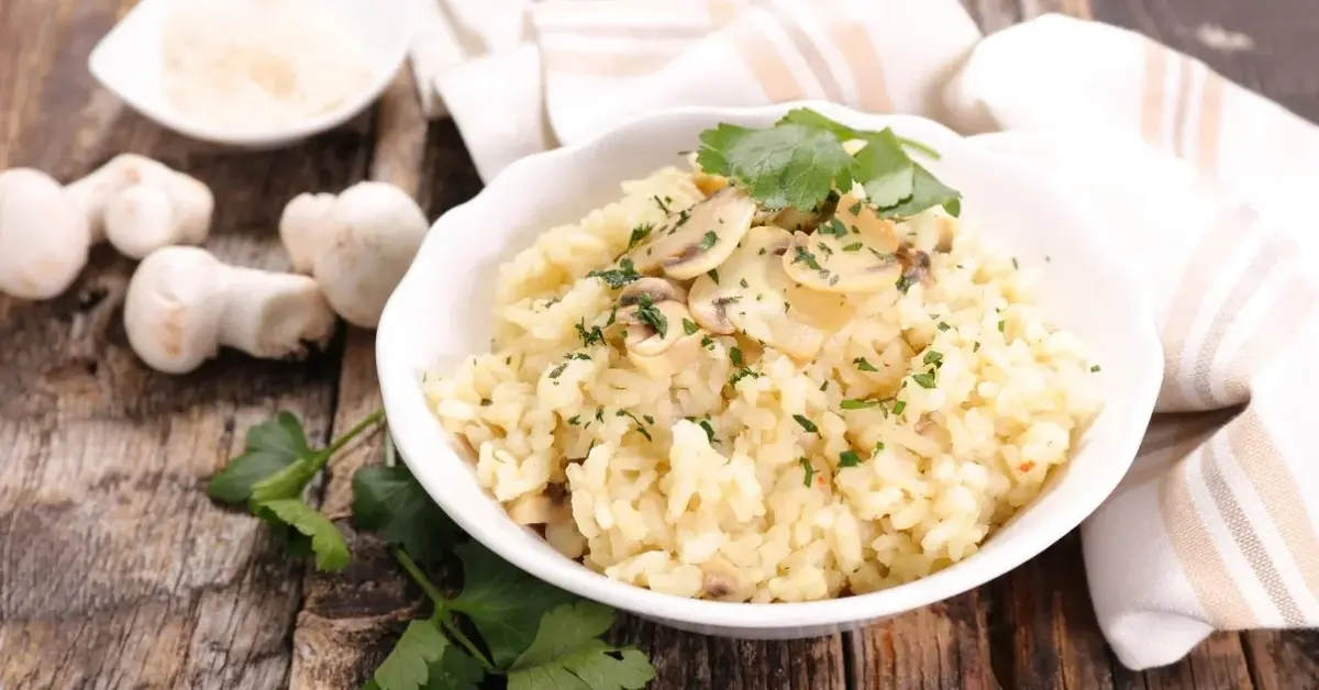 Risotto z pieczarkami w białej miseczce leżącej na drewnianym blacie. Obok biała ściereczka i pieczarki