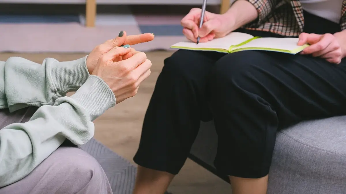 Ręce kobiety siedzącej na krześle, obok kobieta notująca coś na kartce papieru