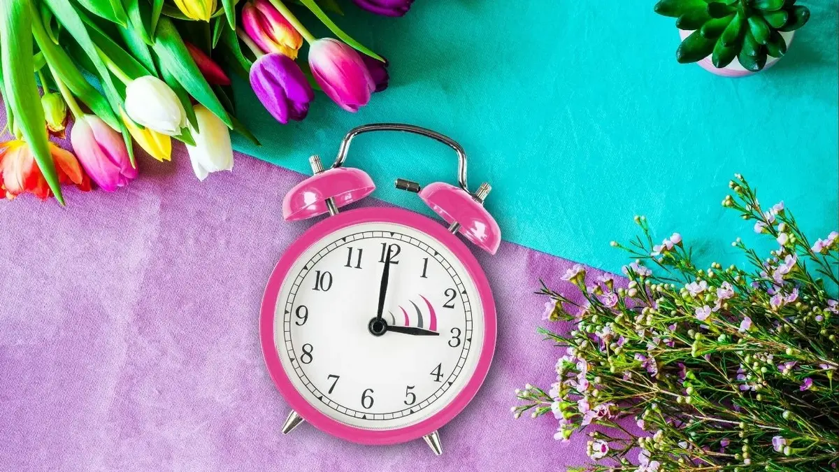 różowy zegar leżący między kwiatami na obrusie niebiesko-fioletowym