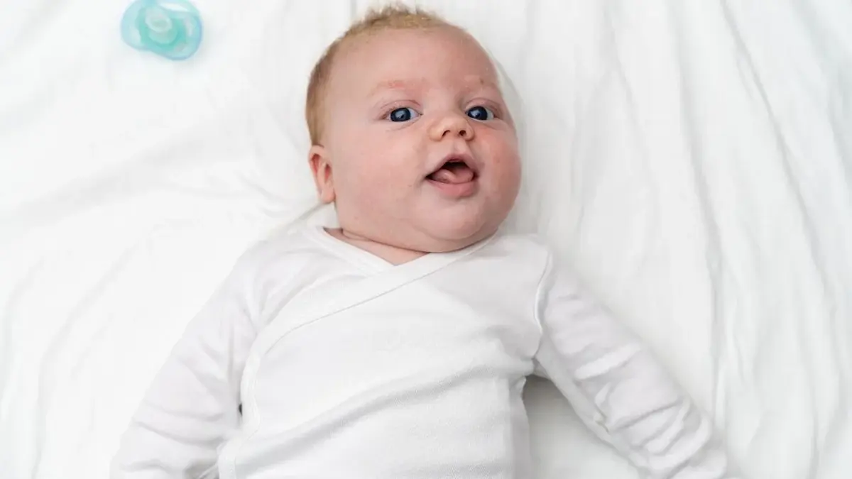 uśmiechnięty noworodek ubrany na biało leży na białej pościeli