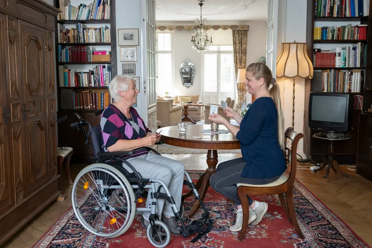 Wnętrze pokoju, przy okrągłym stoliku siedzi starsza kobieta na wózku oraz młodsza blondynka z warkoczem