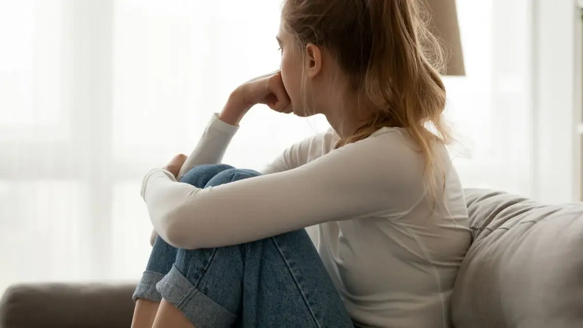 zasmucona kobieta w białej bluzce siedzi na kanapie i patrzy przez okno