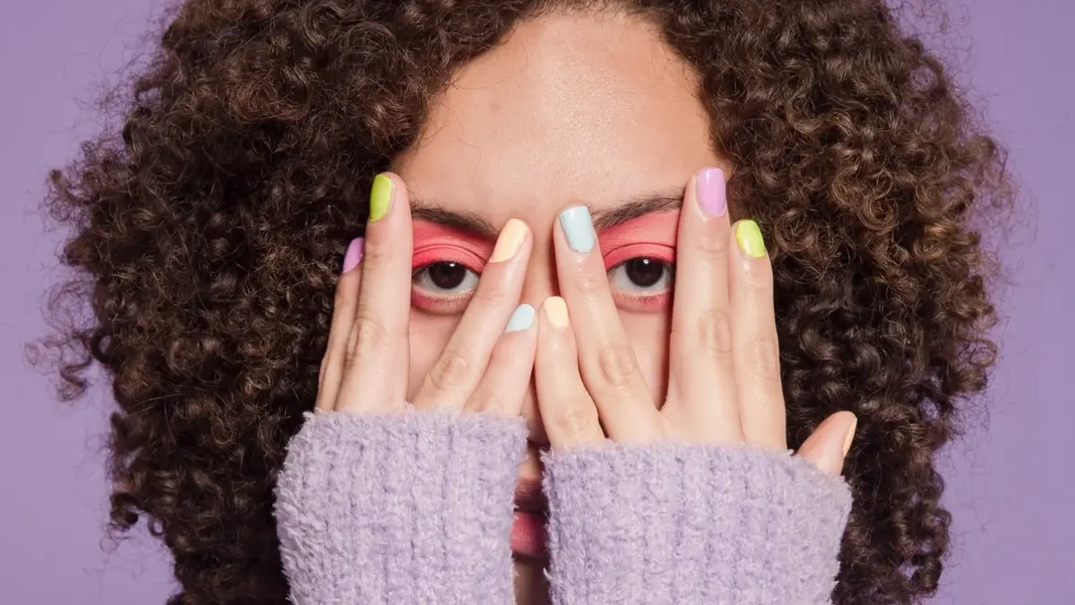 Kobieta o oliwkowej cerze i ciemnych, kręconych włosach przysłania twarz dłońmi o jaskrawie pomalowanych paznokciach
