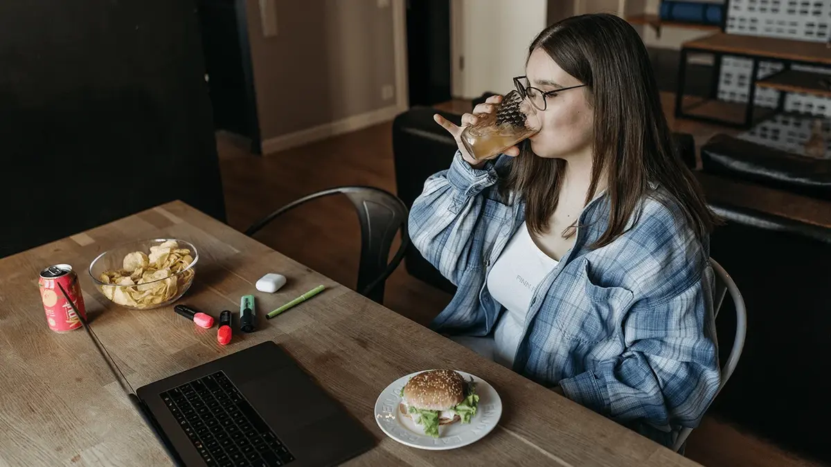 Młoda kobieta w okularach i o długich włosach siedzi przy stole pijąc napój ze szklanki, przednią talerz z kanpką