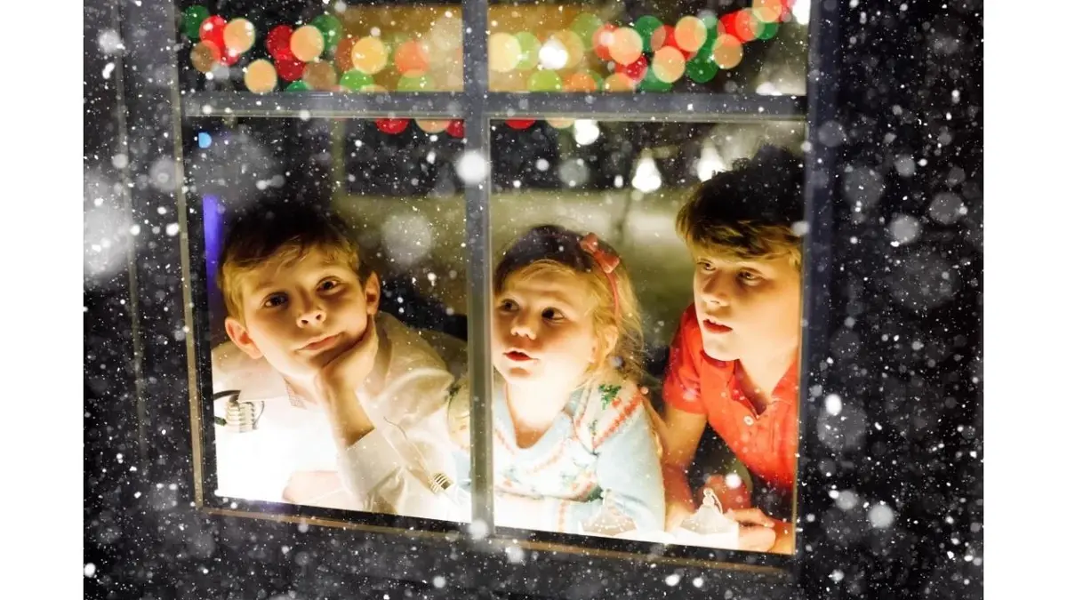 Dzieci patrzą przez oświetlone okno na padający śnieg.