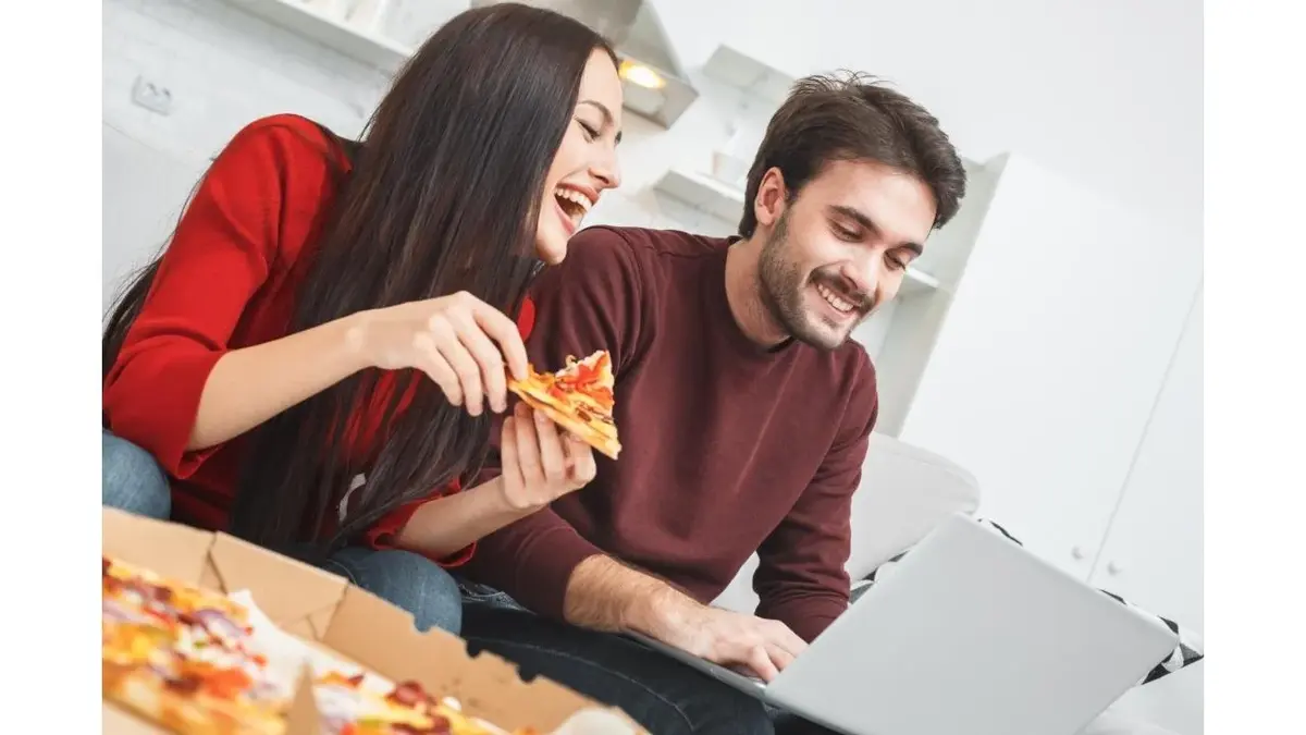 Dziewczyna i chłopak jedzą pizzę, patrzą na laptopa i się śmieją.