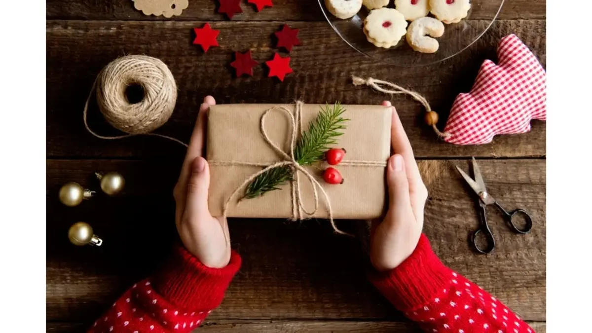 Kobiece ręce trzymają prezent gwiazdkowy. W tel świąteczne ozdoby, sznurek i nożyczki.