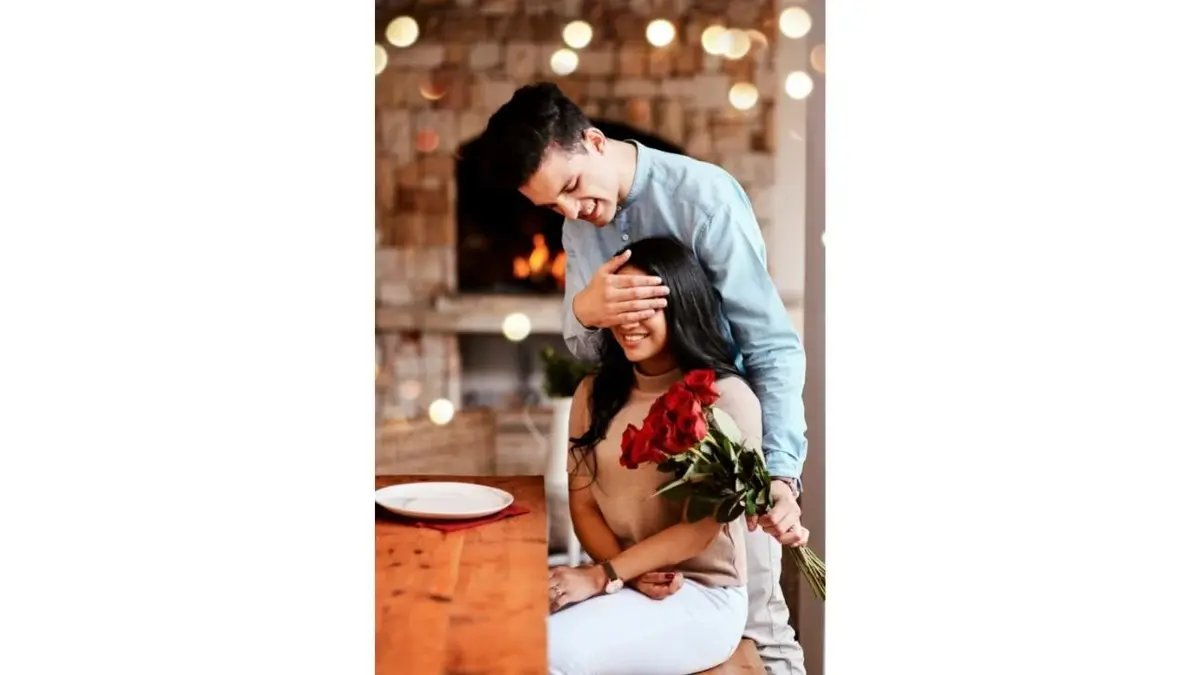 Stojący mężczyzna trzyma róże i zasłania siedzącej kobiecie oczy.