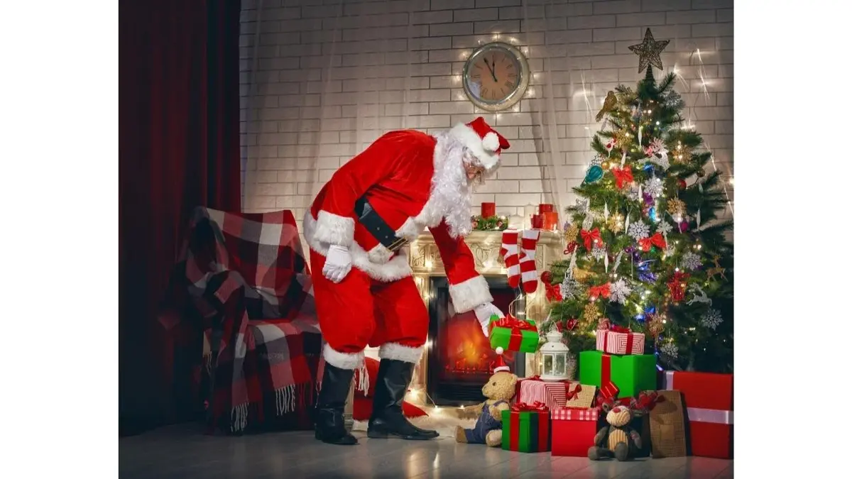 Mikołaj kładzie prezenty pod choinkę. Na podłodze leży dużo prezentów.