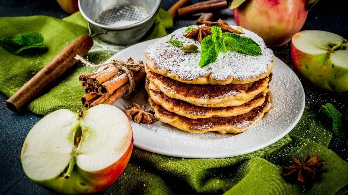 Placuszki z jabłkami posypane cukrem pudrem. Obok jabłka, cynamon.
