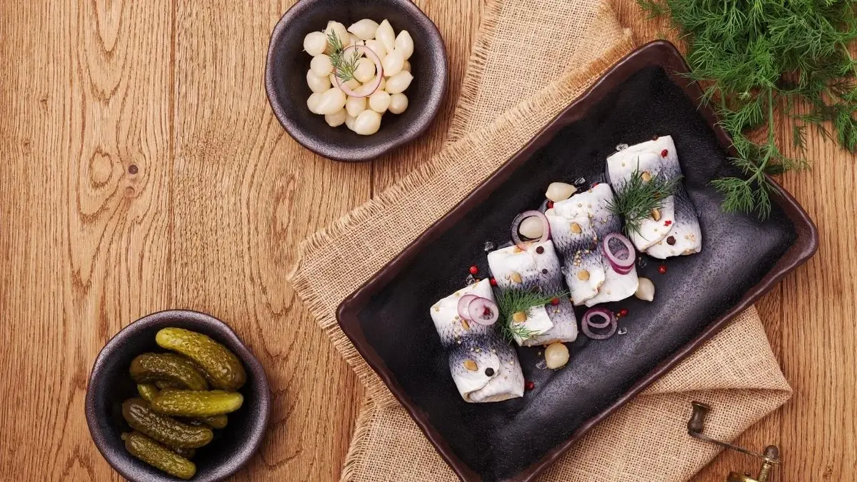 Śledzie z cebulka. Obok leżą ogórki kiszone i szalotki.