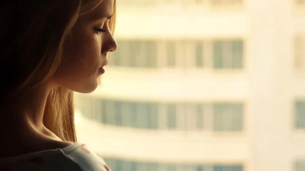 Smutna kobieta o jasnych włosach stoi przy oknie.