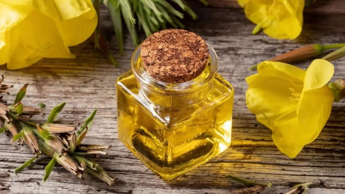 Olej z wiesiołka w karafce. Obok leżą żółte kwiaty wiesiołka.