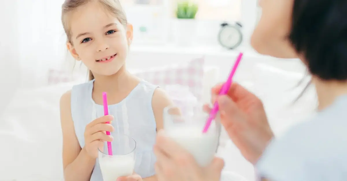 Uśmiechnięta dziewczynka o blond włosach w białej sukience trzymając w ręku szklankę spogląda na pół widoczną mamę