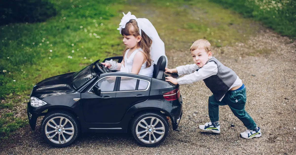 Mały chłopiec pchający samochód zabawkę z dziewczynką w środku, oboje w odświętnych strojach na weselu