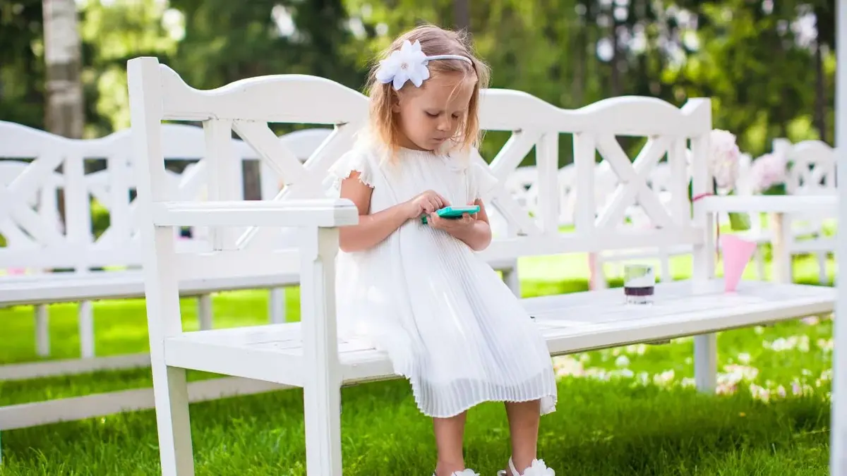 Małą dziewczynka w odświętnej białej sukience siedzi na ławce w trakcie ślubu bawiąc się telefonem