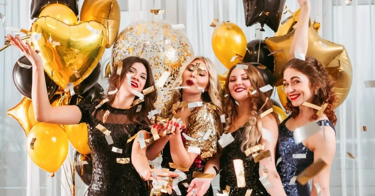 Cztery roześmiane imprezowo ubrane kobiety trzymając dużo czarno złotych balonów bawią się na wieczorze panieńskim