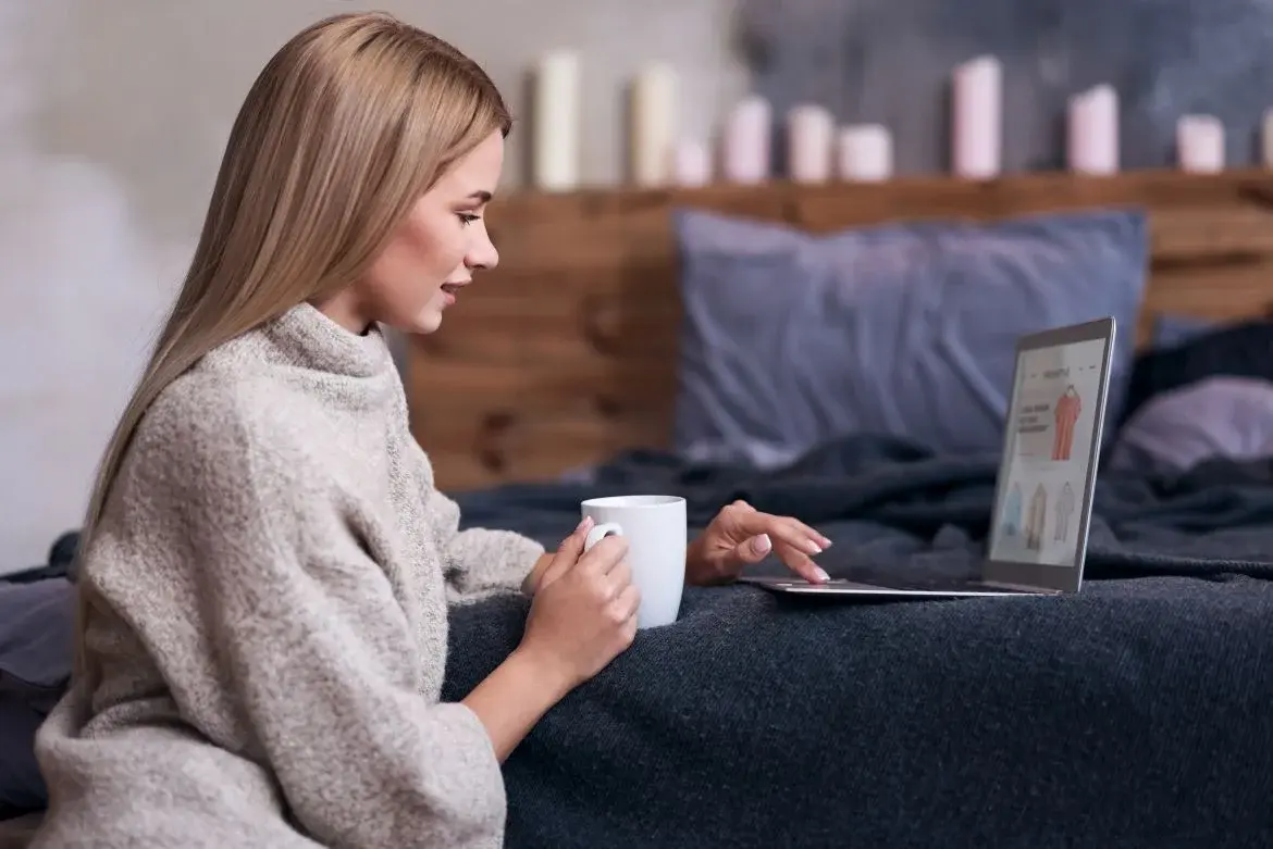 Kobieta długich blond włosach z kubkiem w ręce siedząc w domu robi zakupy online z użyciem cachback
