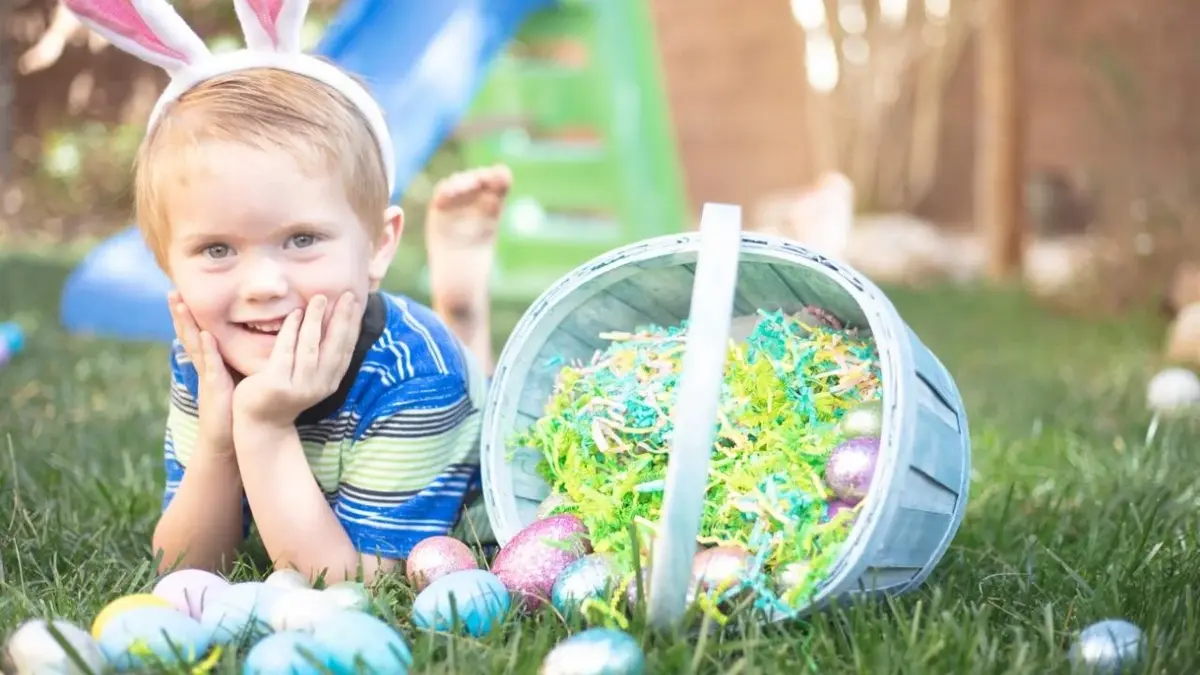 Chłopiec w opasce z uszami zająca, koło koszyka z czekoladowymi jajkami wielkanocnymi..