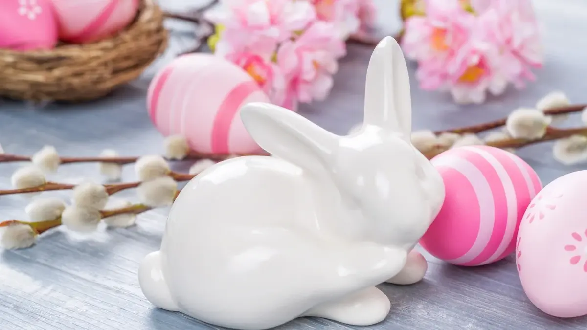 Dekoracje na Wielkanoc: biały porcelanowy króliczek, bazie, różowe jajka i różowe kwiaty.